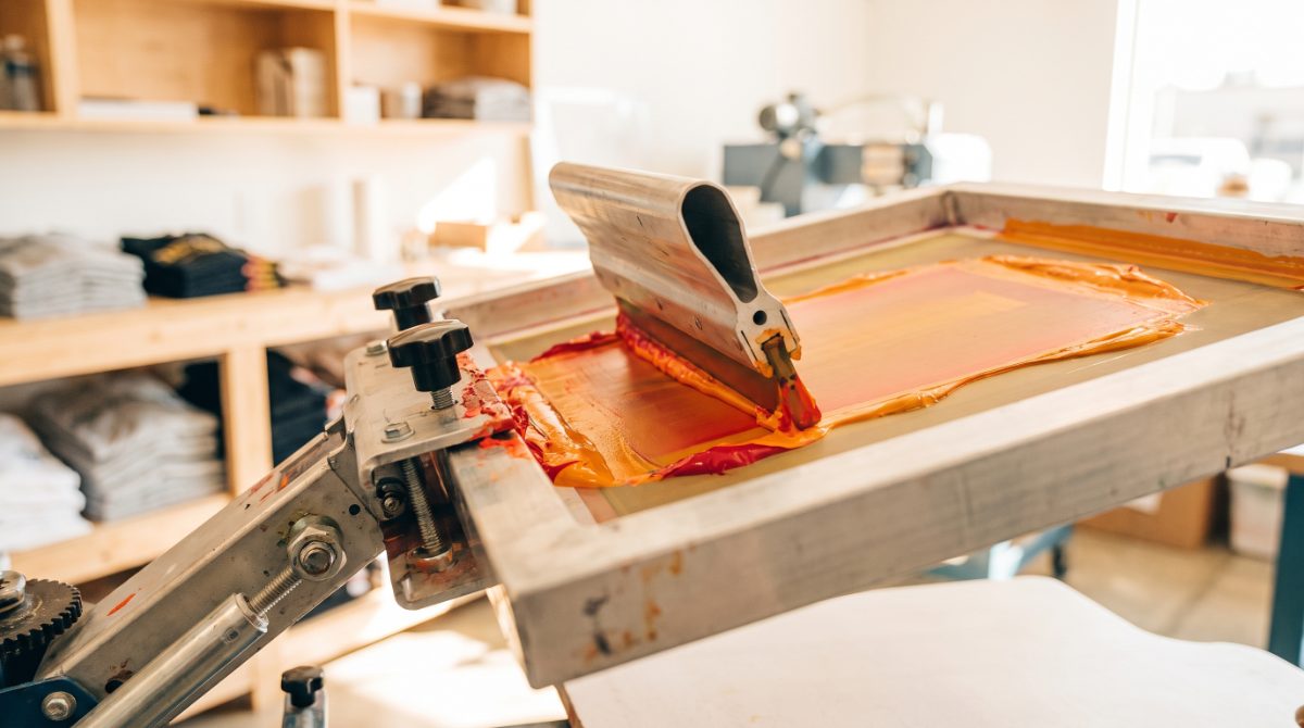 Squeegee pulling vivid ink across a screen printing mesh frame on an automatic press